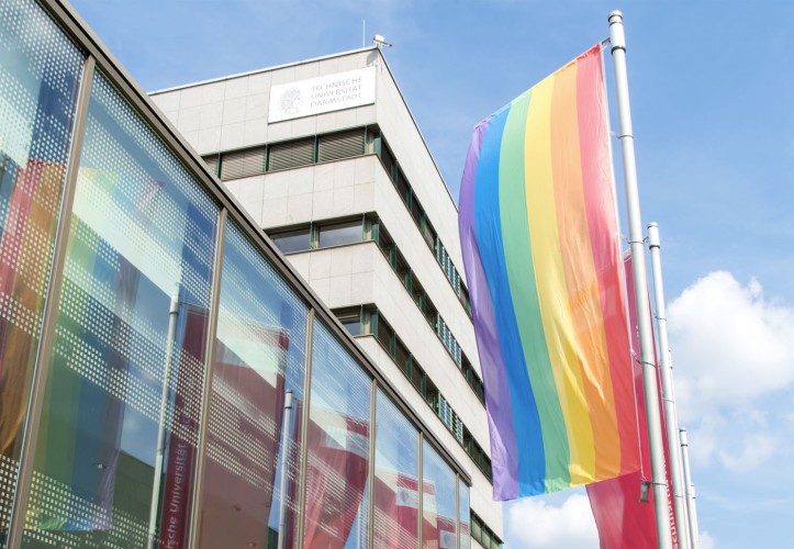 Das Bild zeigt eine wehende Regenbogenflagge vor dem TU Gebäude Karo 5. Die Fahne spiegelt sich in den Fenstern, darüber ist der blaue Himmel.