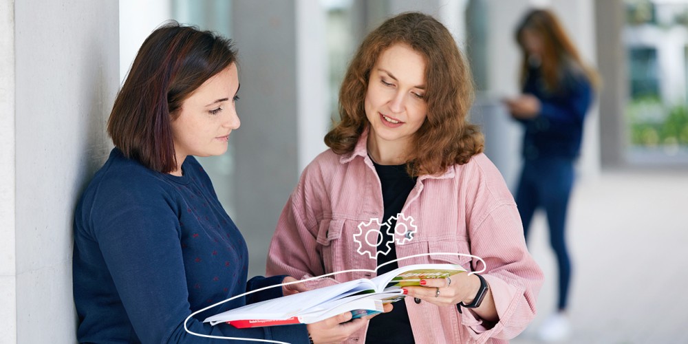 Zwei Frauen stehen draußen an Säulen angelehnt und schauen gemeinsam in ein Arbeitsbuch