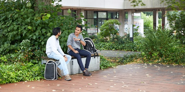 Zwei Männer sitzen draußen auf einer Bank, im Hintergrund grüne Vegetation und ein Gebäude