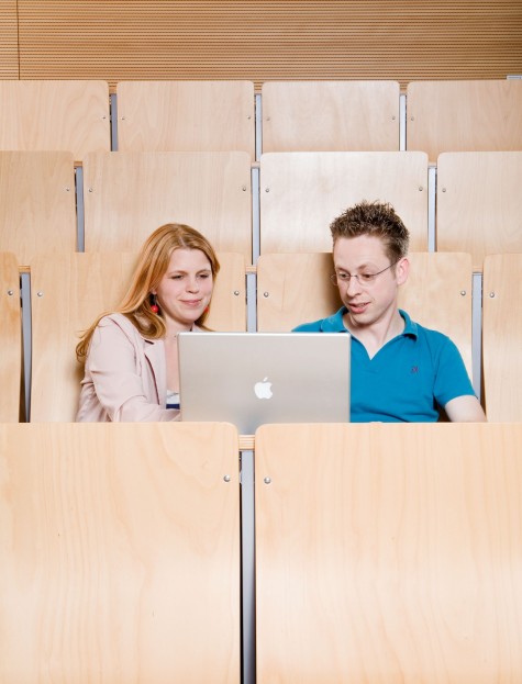 Eine wissenschaftliche Mitarbeiterin und ein wissenschaftlicher Mitarbeiter sitzen vor Laptop in Hörsaalbestuhlung