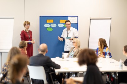 Zwei Referenten stehen vor einem Flipchart. Die Workshopteilnehmenden sitzen mit konzentriertem Gesichtsausdruck um Tische und sammeln Begriffe.